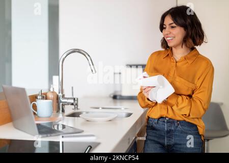 Donna araba che si asciuga le mani dopo aver lavato i piatti e usando il laptop in cucina Foto Stock