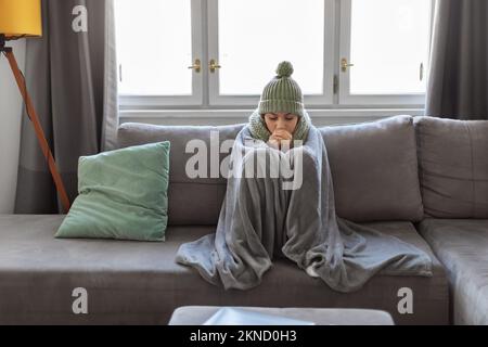 Donna araba che soffre freddo a casa, seduta sul divano coperto di coperta Foto Stock