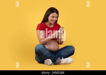 Felice donna incinta che usa lo smartphone mentre si siede sul pavimento su sfondo giallo Foto Stock