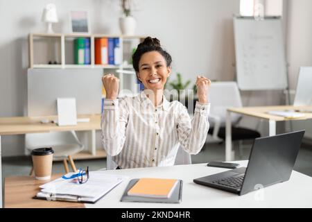 Fortuna per il business. Un'imprenditrice femminile molto gioita che usa il laptop e che scuote i pugni di gioia, festeggia grandi notizie, seduto in ufficio Foto Stock