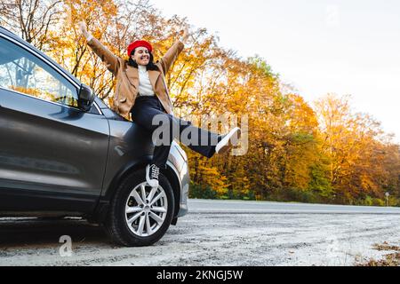 una donna elegantemente vestita siede sul cappuccio di un'auto con le braccia sollevate Foto Stock