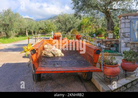 Trattore e rimorchio con rastrello e potatrice di olive parcheggiate nel villaggio greco con oliveti sul fianco della collina e montagne blu e nebbia sullo sfondo Foto Stock