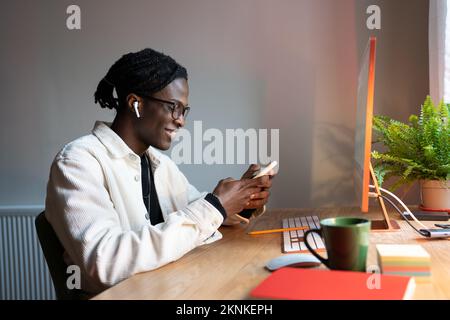 Giovane studente africano che usa lo smartphone, chattando con gli amici mentre studia online a casa Foto Stock