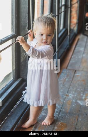 bellissimo bambino biondo in una nightgown vicino alla finestra Foto Stock