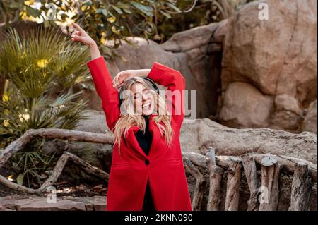 Una donna allegra dai capelli leali in un cappotto rosso che suona musica con le cuffie in un parco Foto Stock