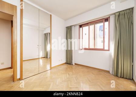 Camera da letto vuota con armadio a muro con porte a specchio e bagagliaio, finestra in alluminio anodizzato rosso e tende con teli, legno di pino sul Foto Stock