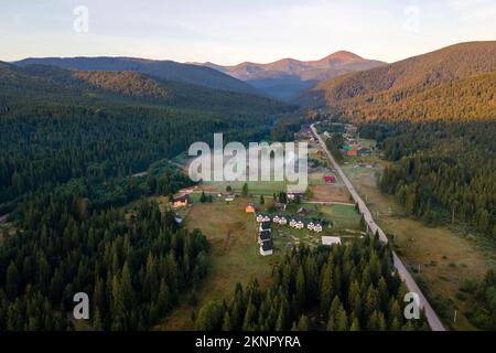 Vista aerea della serata nebbia sulle piccole case di un villaggio di montagna tra alte vette con alberi di pino scuro al tramonto luminoso. Amazingl scenario di Foto Stock