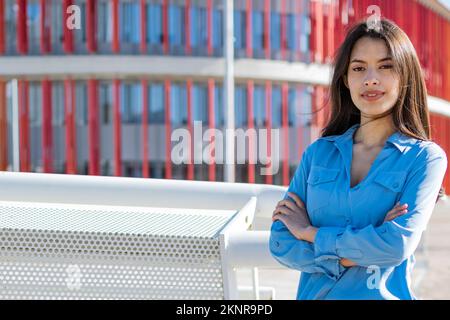 donna d'affari in posa fiduciosa con le braccia incrociate con rosso e vetro moderno edificio ufficio sullo sfondo Foto Stock