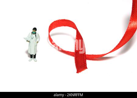 Fotografia di giocattoli di figura della gente in miniatura. Giornata mondiale dell'AIDS concetto di giornata di consapevolezza. Ragazza donna medico in piedi accanto al nastro rosso. Isolato su fondo bianco gr Foto Stock