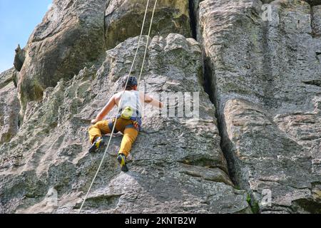 Giovane uomo che sale ripida parete di montagna rocciosa. Il climber maschile supera il percorso impegnativo. Il concetto di sport estremi Foto Stock