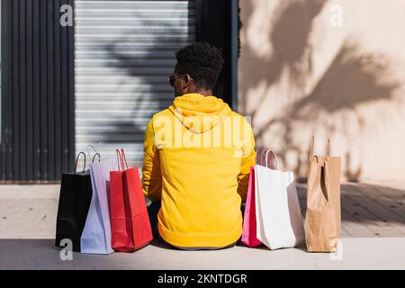 Un giovane uomo africano vestito con una felpa gialla seduta su una panca di strada accanto ad alcune borse della spesa visto dalla sua schiena. Foto Stock