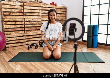 Giovane donna ispanica registrazione tutorial di allenamento in palestra guardando positivo e felice in piedi e sorridente con un sorriso fiducioso mostrando i denti Foto Stock