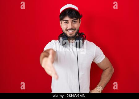 Uomo ispanico con barba che indossa cappello da giocatore e cuffie sorridente amichevole offrendo handshake come saluto e benvenuto. business di successo. Foto Stock