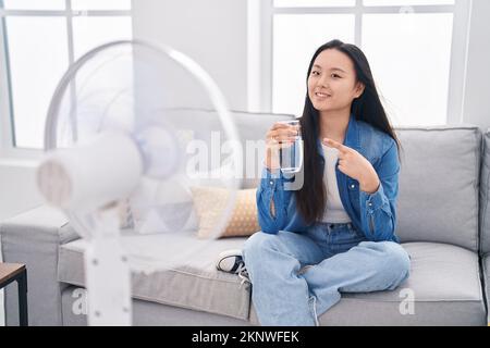 Giovane donna asiatica bere un bicchiere d'acqua godendo aria dal ventilatore sorridendo felice indicando con mano e dito Foto Stock