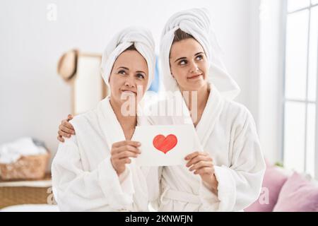 Donna e figlia di mezza età che indossano un accappatoio che tiene la scheda del cuore sorridente guardando al lato e fissando via pensando. Foto Stock