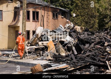 Pulisca-in su il lavoratore vicino alla casa danneggiata di stile del cottage del fuoco con i residui di un edificio commerciale bruciato. Foto Stock