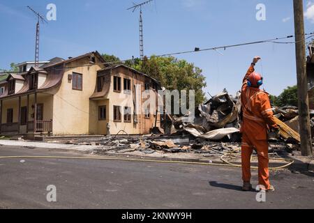 Pulisca-in su il lavoratore vicino alla casa danneggiata di stile del cottage del fuoco con i residui di un edificio commerciale bruciato. Foto Stock