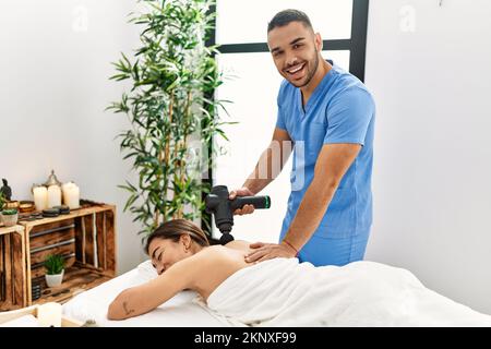 Uomo latino e donna che indossano l'uniforme di fisioterapia massaggiante indietro usando la pistola a percussione al centro di bellezza Foto Stock