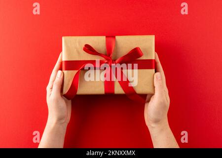 Testa di mani che tiene il regalo in carta marrone con nastro rosso, su sfondo rosso con spazio di copia Foto Stock