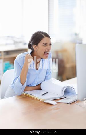 Quel momento in cui scoprite il vostro lavoro duro ha pagato fuori. Una donna d'affari eccitata che fa una pompa di pugno mentre si siede alla sua scrivania dell'ufficio. Foto Stock