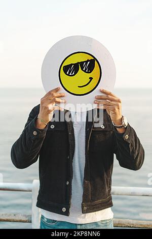 Giovane uomo adulto faccia coperta con un sorriso emoji viso con occhiali da sole, disegnata a mano su un disco bianco in piedi all'aperto contro il mare-social network in Foto Stock