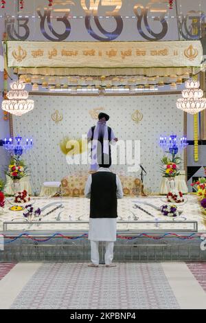 Un sikh pries (primo piano) conduce un servizio mentre un secondo sacerdote ondeggia il suo flan giallo cerimoniale sulla bibbia sikh. . In un tempio di Queens, New York Foto Stock
