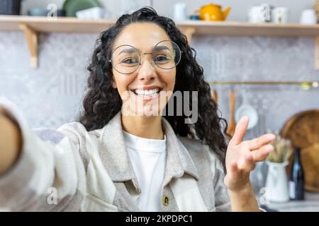 Primo piano. Giovane bella donna latino-americana che parla in videochiamata a casa, blogging, prendendo selfie, sorridendo. Ondeggiando alla fotocamera. Foto Stock