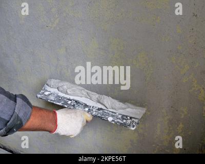 La mano del costruttore in un guanto tiene una spatola larga con un mortaio di cemento spesso vicino ad una parete innescata con uno spazio vuoto per il testo, lavori di intonaco sulla parete Foto Stock