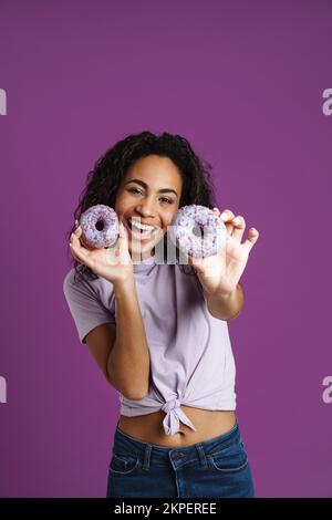 Giovane donna nera che ride mentre si posa con le ciambelle isolate su sfondo viola Foto Stock