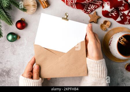 Donna mani in maglione tenendo carta e busta bianca. Piatto di sfondo grigio con tazza di caffè e decorazione natalizia. Vista dall'alto Foto Stock