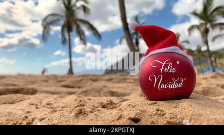 Natale bomba nel cappello di Babbo Natale con parole buon Natale in spagnolo sulla spiaggia distesa sulla sabbia con palme e cielo blu sullo sfondo. Foto Stock