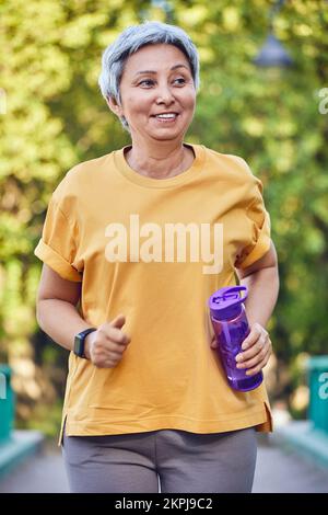 Donna asiatica in forma matura in abbigliamento sportivo tenendo una bottiglia d'acqua fare esercizio all'aperto, jogging nel parco estivo. Stile di vita attivo e sano dei pensionati, Foto Stock