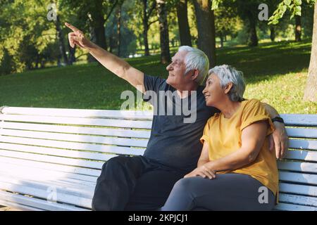 Positivo multi razziale anziani 60s coppia che riposano sedersi sulla panca nel parco dopo una passeggiata, marito mostrando punta dito in su parlare con sorridente moglie rilassante o Foto Stock