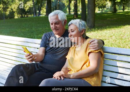 Sposi maturi siedono sulla panchina nel parco estivo con smartphone. Coppia con capelli grigi guardare lo schermo del cellulare godere di una nuova applicazione all'aperto utilizzare la tecnologia moderna Foto Stock