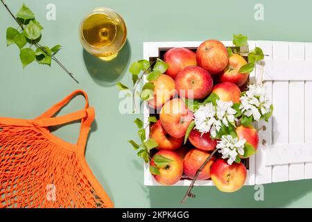 Composizione ecologica con mele da fattoria in una scatola di legno bianco con rami in fiore, bicchiere con succo di mela naturale e sacco di cotone su dorso verde Foto Stock