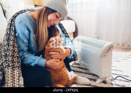 Uso del riscaldatore a casa in inverno freddo. Donna che gioca con il gatto con un dispositivo elettrico indossando un maglione e un cappello sotto coperta. Sentiti accogliente con gli animali domestici Foto Stock