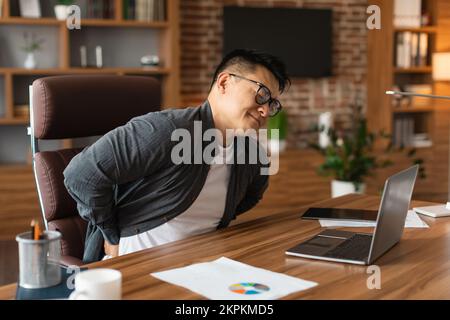 Infelice uomo asiatico di mezza età manager in occhiali che soffrono di dolori muscolari alla schiena a tavolo con notebook Foto Stock