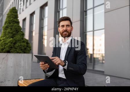 Nuova app, annuncio e offerta. Un giovane uomo europeo sorridente con un tablet per l'uso della barba in tuta, si siede sulla panca Foto Stock