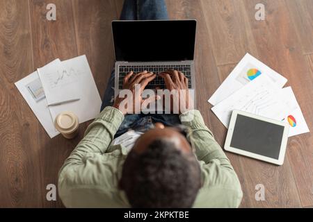 Uomo afro-americano di mezza età in occhiali che scrivono sul laptop con lo schermo vuoto siede sul pavimento con i grafici Foto Stock