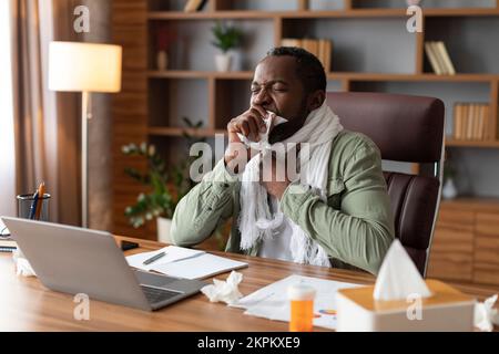 Triste uomo d'affari afro-americano di mezza età in tosse sciarpa e che soffre di malattia a tavola con il portatile Foto Stock
