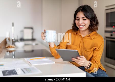 Donna araba sorridente che beve caffè e usa un tablet digitale in cucina Foto Stock