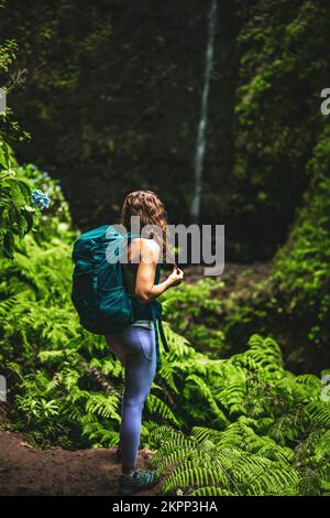 Descrizione: Donna sportiva con zaino si meraviglia in una cascata impressionante nella giungla con bellissimi fiori e felci. Levada, di Caldeirão Verde, Foto Stock