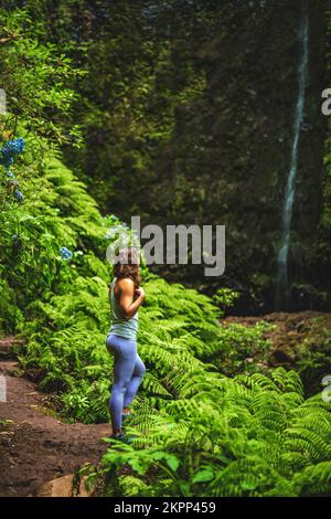 Descrizione: Donna sportiva si meraviglia in una cascata impressionante nella giungla con bellissimi fiori e felci. Levada di Caldeirão Verde, Isola di Madeira Foto Stock