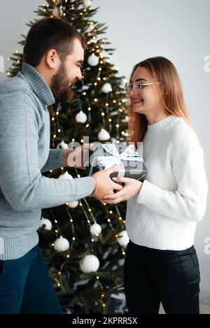 Un giovane e una ragazza che si amano abbracciano e baciano in maglioni accoglienti vicino a un piccolo albero di Natale Foto Stock