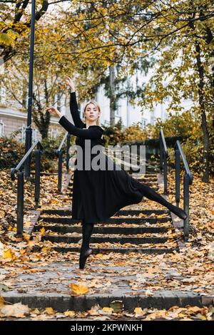 Ballerina Dancing in via autunnale città, ballerino moderno in vestito nero, Pointe Shoes Outdoors. Balsamo, balsamo quotidiano Foto Stock