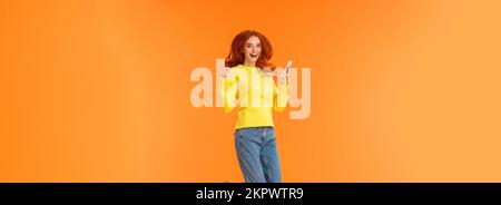 Verticale a tutta lunghezza shot spensierato, lieti di bella ragazza di zenzero in maglione di lana, jeans, saltando su sfondo arancione, tenendo lo smartphone Foto Stock