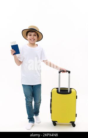 Ragazzo viaggiatore adolescente in t-shirt casual, jeans e cappello di paglia, passeggiate con carta d'imbarco e valigia su sfondo bianco. Foto Stock