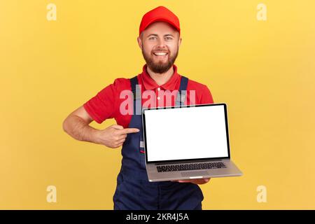 Felice lavoratore uomo in piedi uniforme e puntando lo schermo vuoto del notebook, l'area della pubblicità, guardando la fotocamera con un sorriso toothy. Studio al coperto isolato su sfondo giallo. Foto Stock