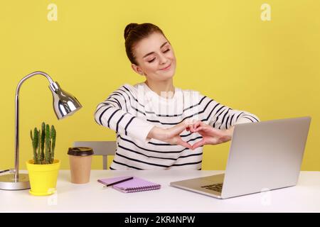 Donna positiva che mostra un gesto cardiaco con le mani sullo schermo del notebook seduto sul posto di lavoro con il notebook, sorridente esprimendo sentimenti amorosi e romantici. Studio interno girato isolato su sfondo giallo Foto Stock