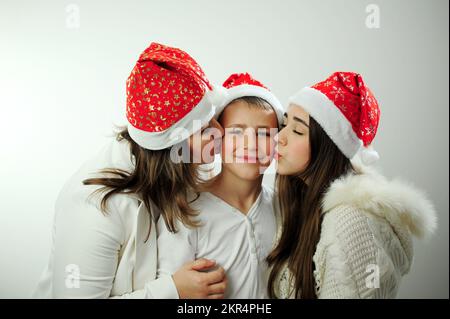 Mamma figlia e figlio celebrare il nuovo anno 3 persone sono vestiti in bianco, stanno indossando cappelli rossi di nuovo anno famiglia incompleta abbracciando Kissing mamma e figlia due lati del loro figlio e fratello Foto Stock